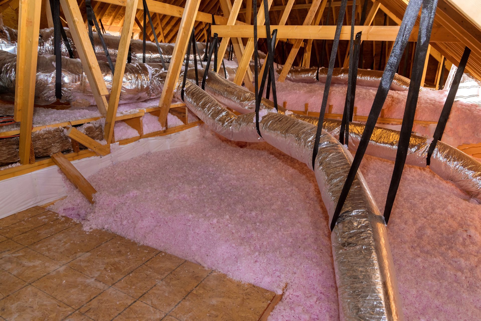 Attic with pink insulation and silver ductwork suspended from wooden rafters.