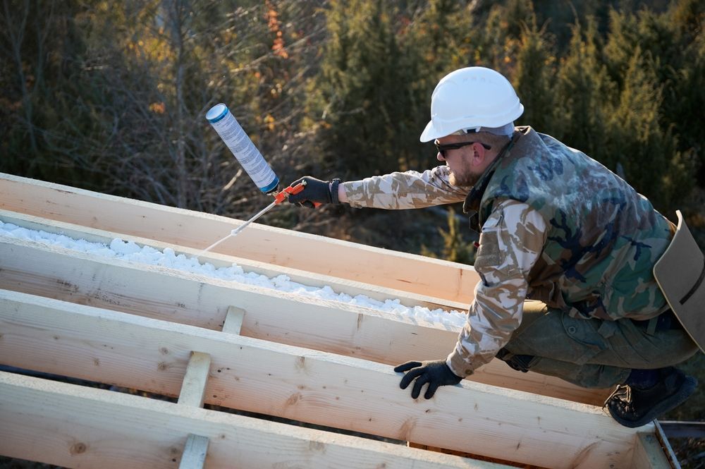 Construction worker in camo using foam sealant on wooden beams outdoors.