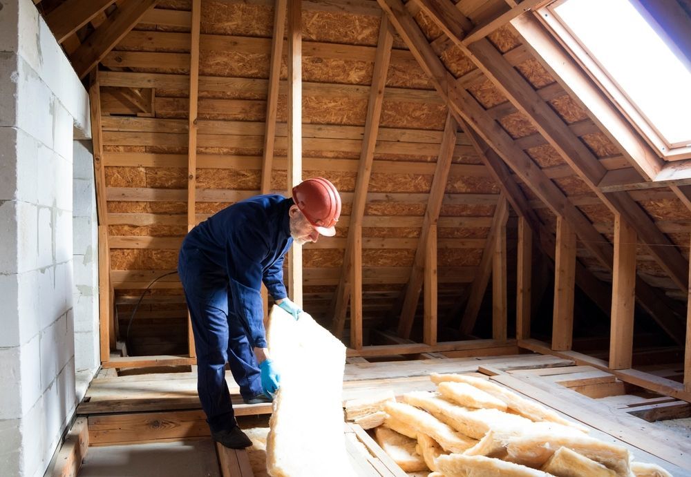 Person in blue workwear installing insulation in an attic, with wooden beams and skylight.