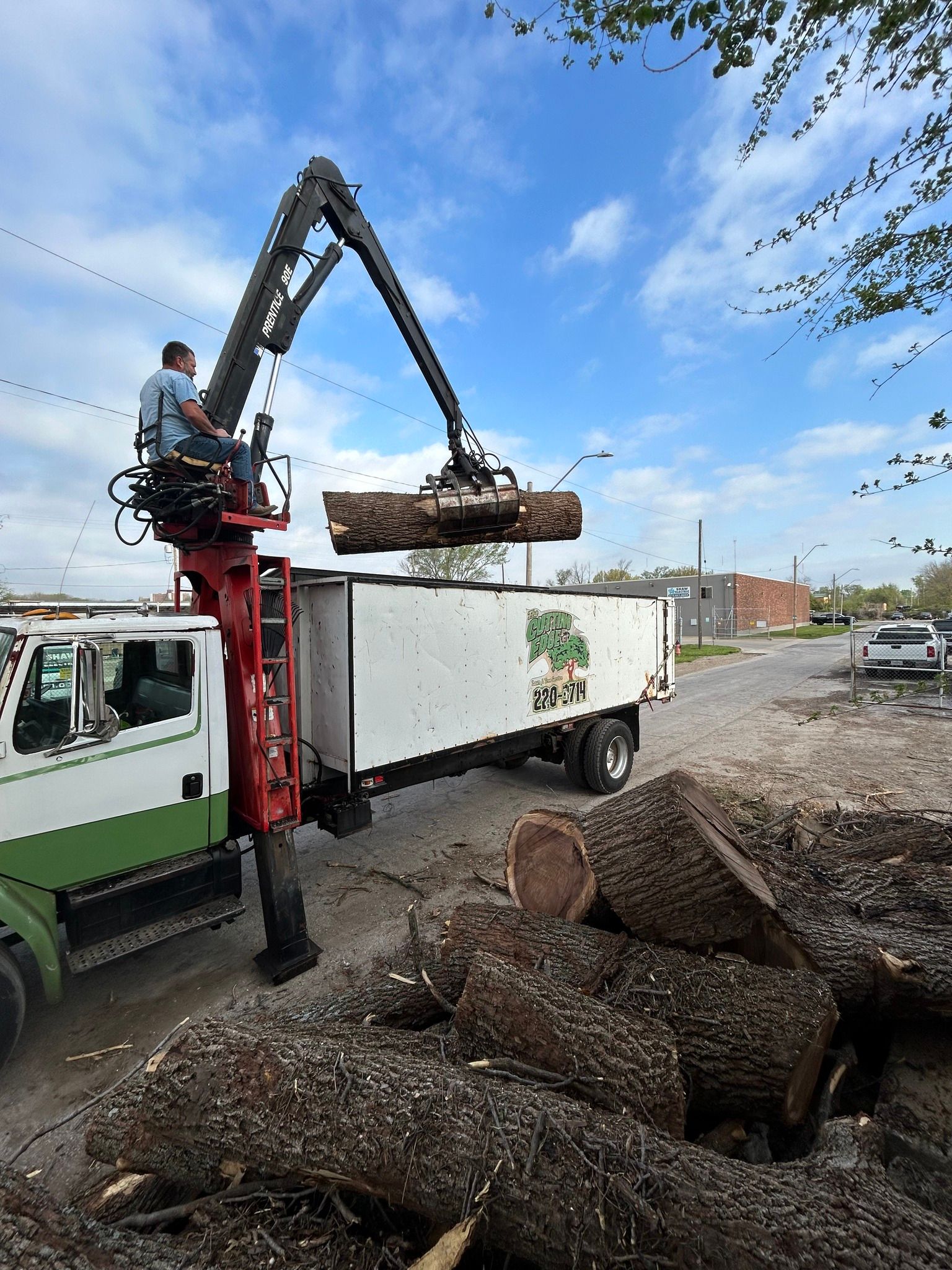 Truck with crane loading a log into a container, with a man operating the crane. Logs on the ground.