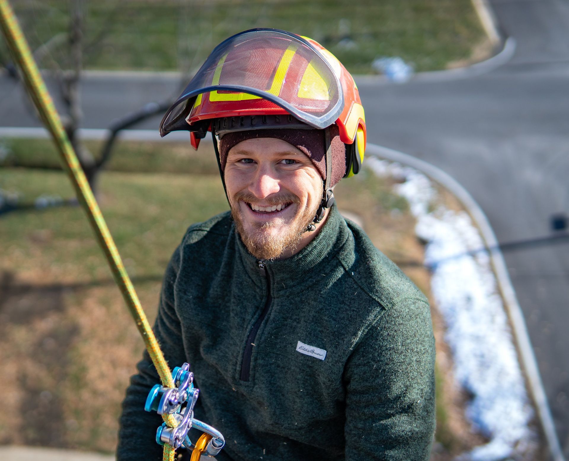 Smiling person in safety helmet and green sweater on a rope. Outdoor setting with road and snow.