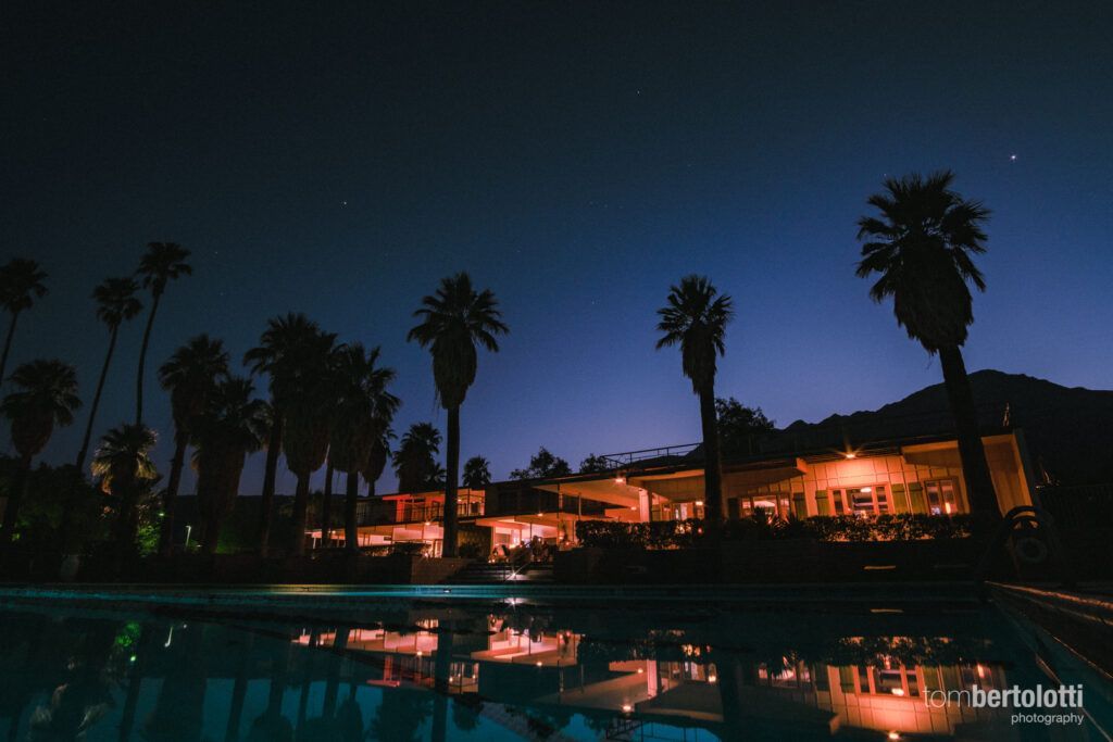 A swimming pool with palm trees in the foreground and a house in the background at night.