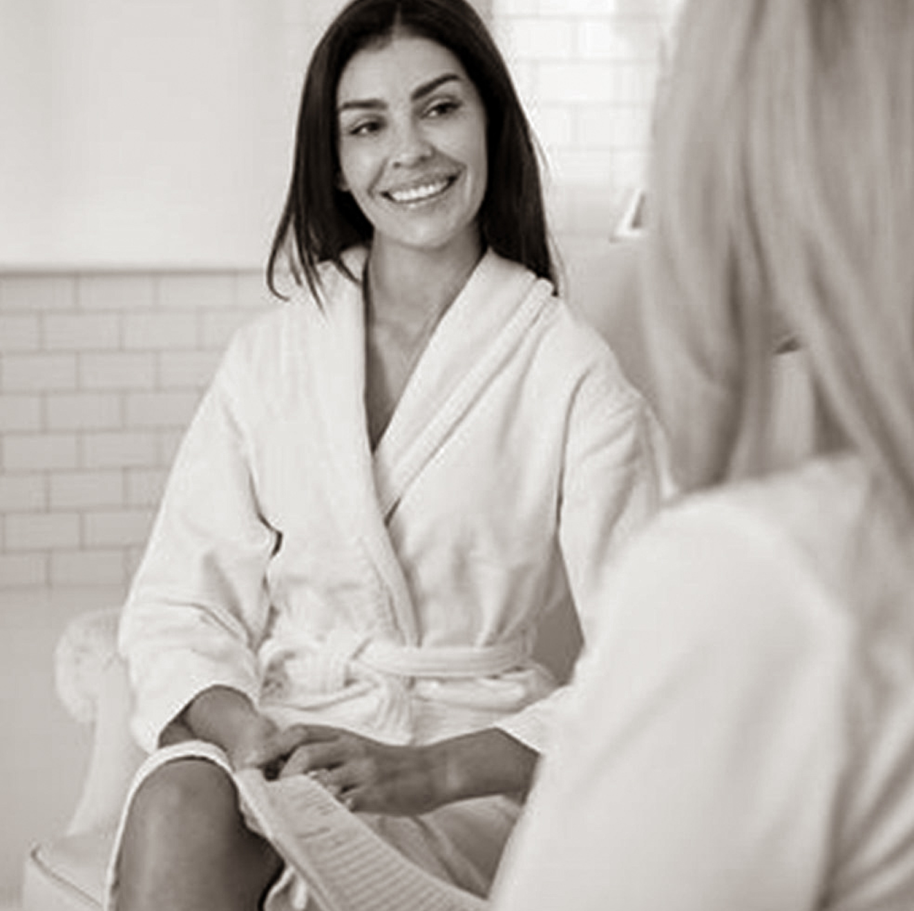 A woman in a bathrobe is sitting next to another woman in a bathtub.