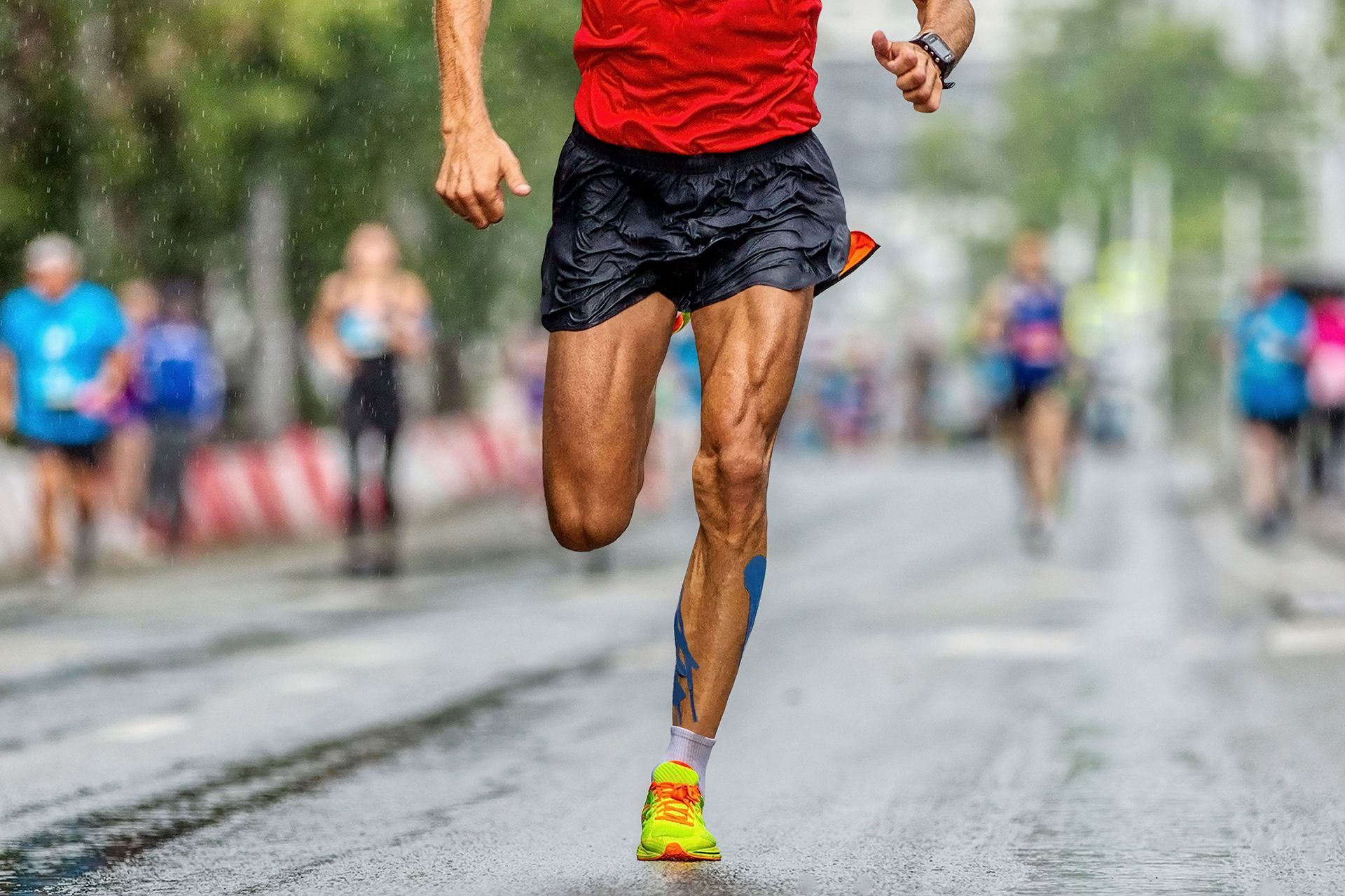 A runner in a red shirt and black shorts sprints along a wet city street, with blurred figures in the background.