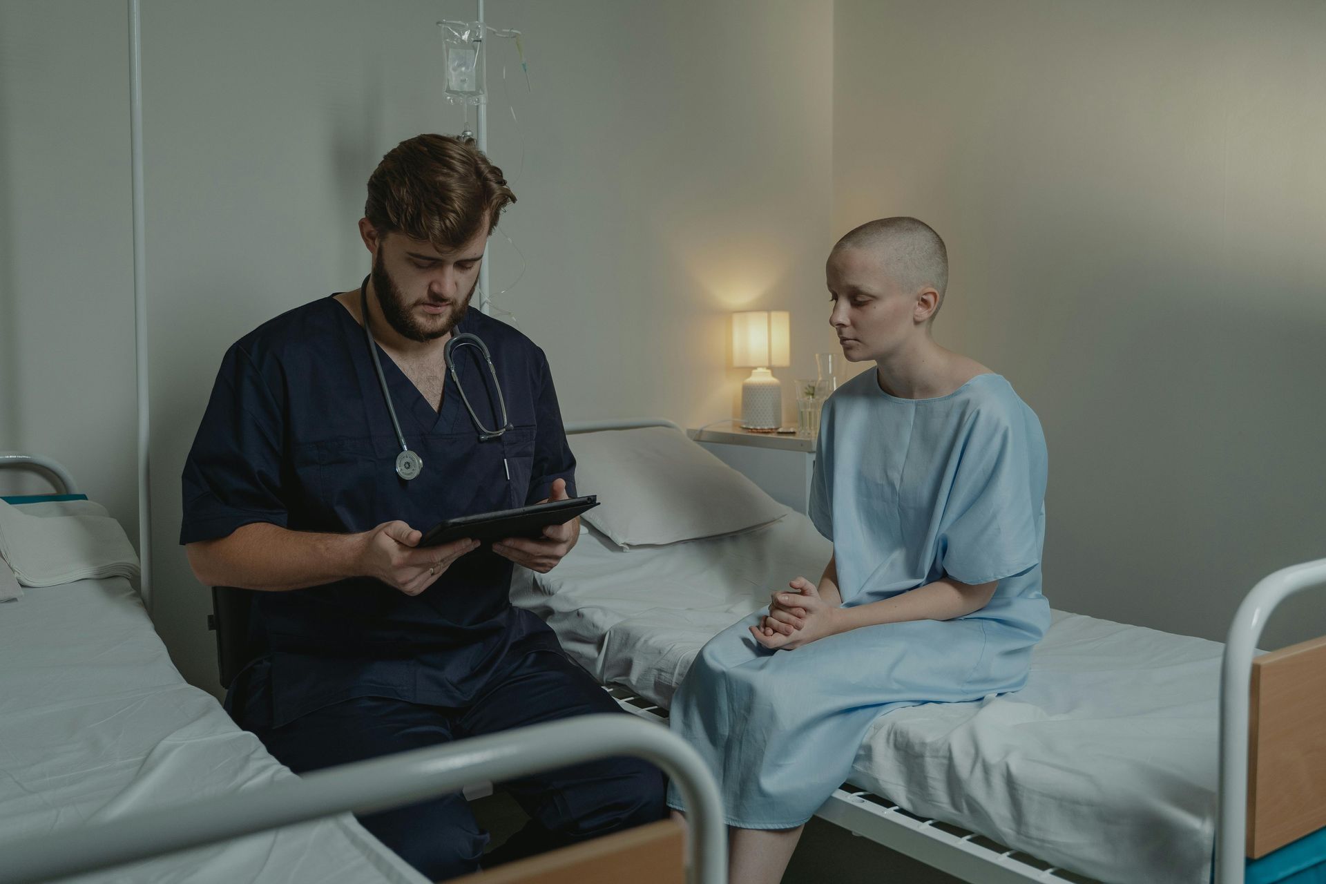 Doctor in blue scrubs consults with a bald patient in a hospital room.