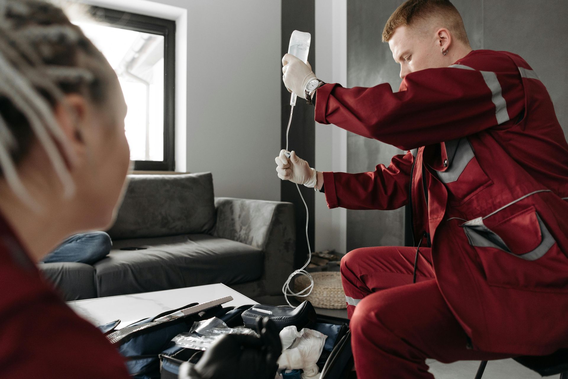 Paramedic in red uniform prepares IV bag indoors near another person.