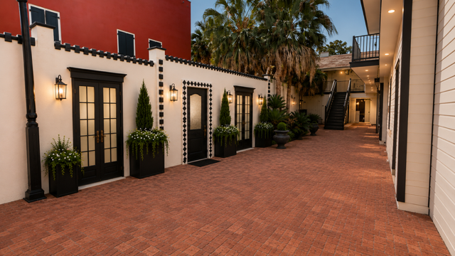 Brick-paved courtyard with Spanish-style buildings, black-trimmed doors, potted plants, and warm evening lighting