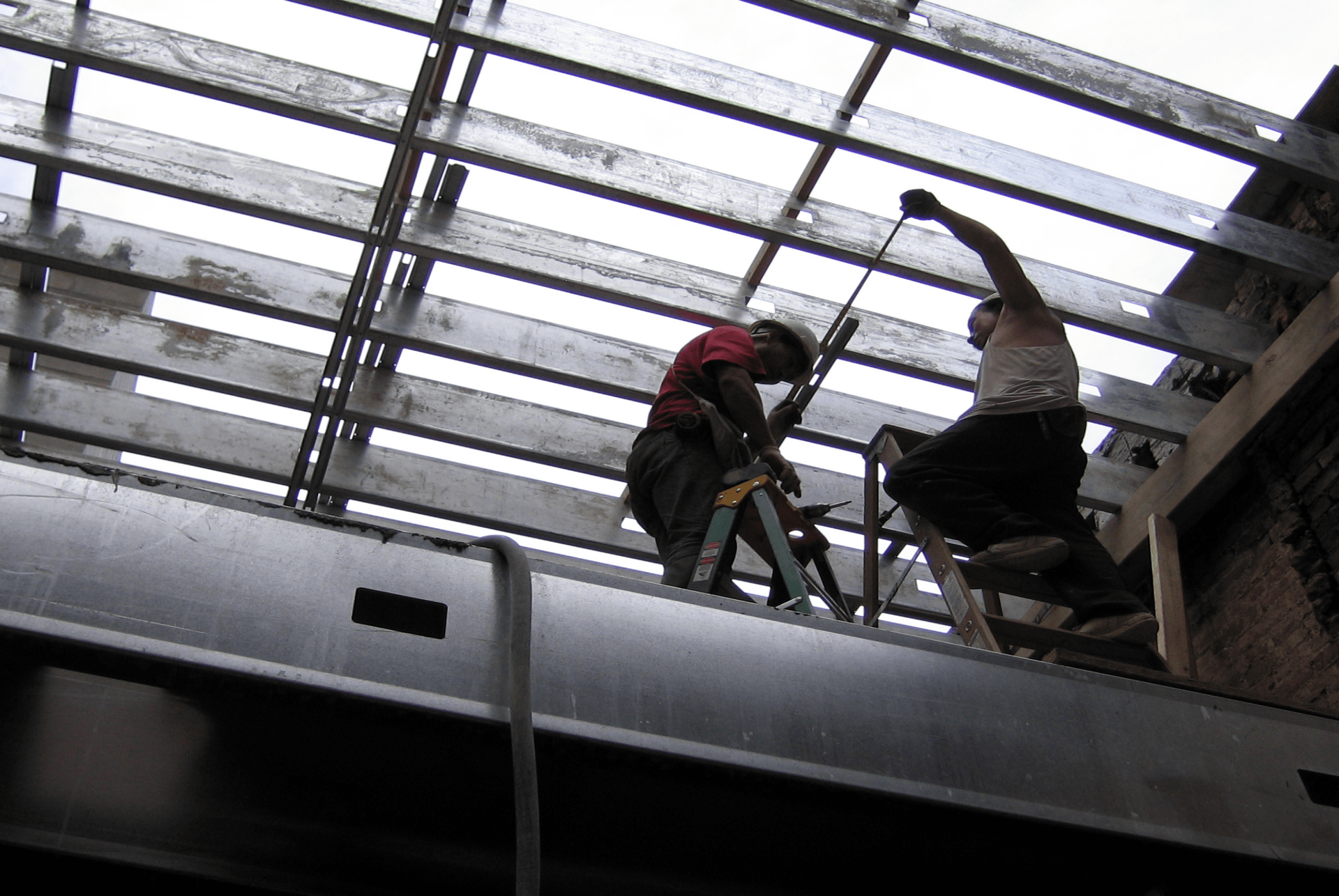 Two construction workers on a steel framework, measuring and working. Overcast sky visible above.