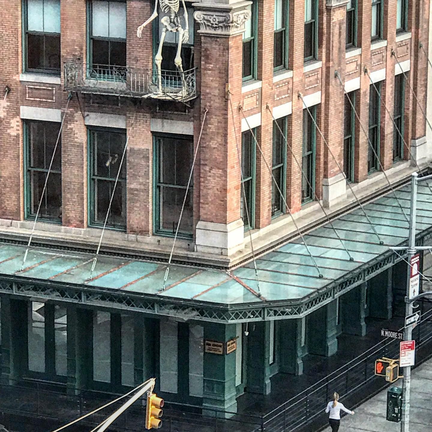 Brick building with green awning, fire escape, skeleton decoration. Pedestrian crossing street, traffic light visible.
