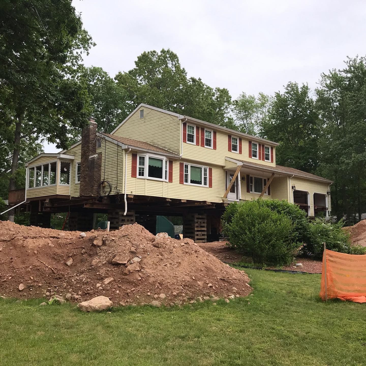 Yellow house raised on supports during construction, surrounded by dirt piles and grass.