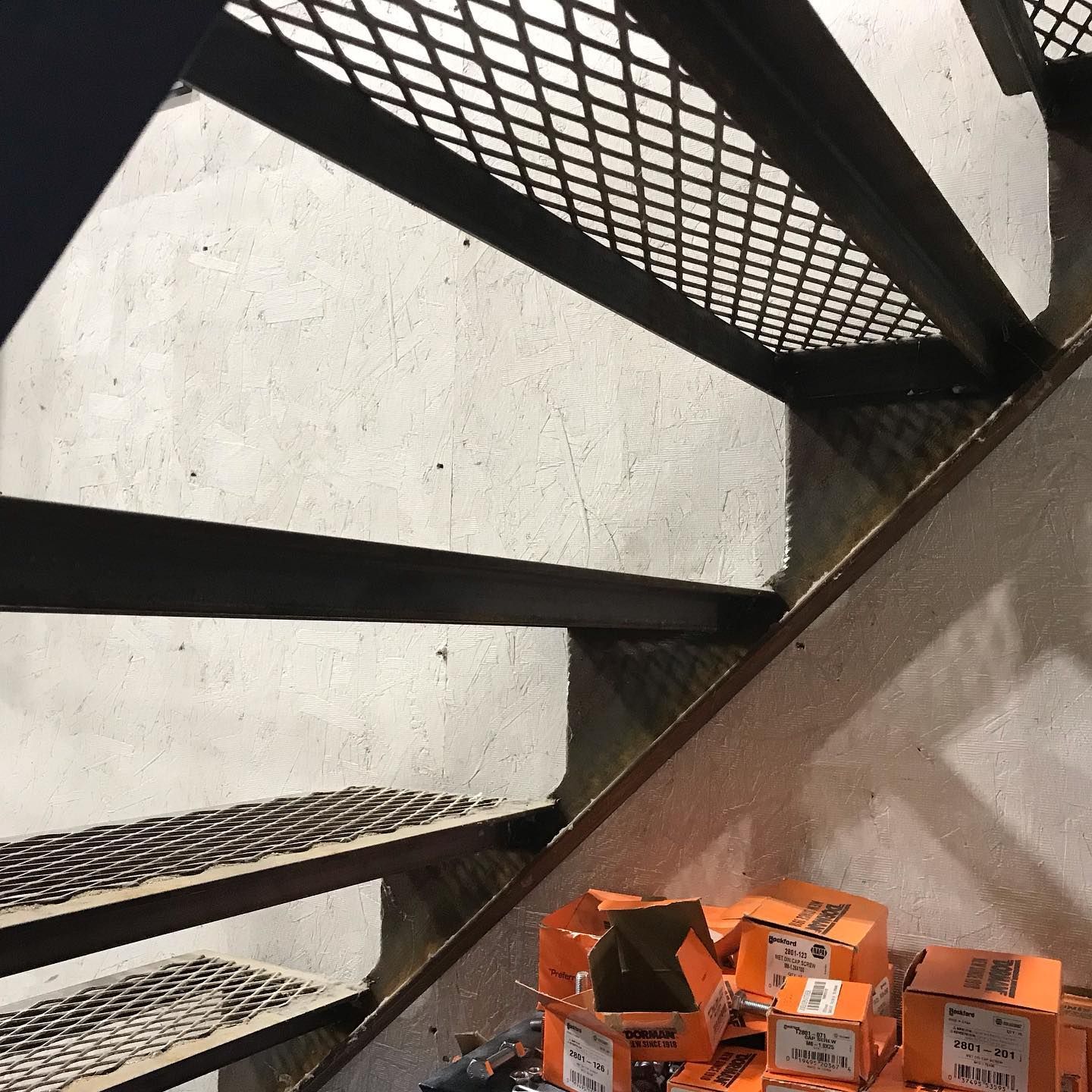 Metal staircase with open-grid treads, casting shadows on a concrete wall. Boxes are stacked at the base.