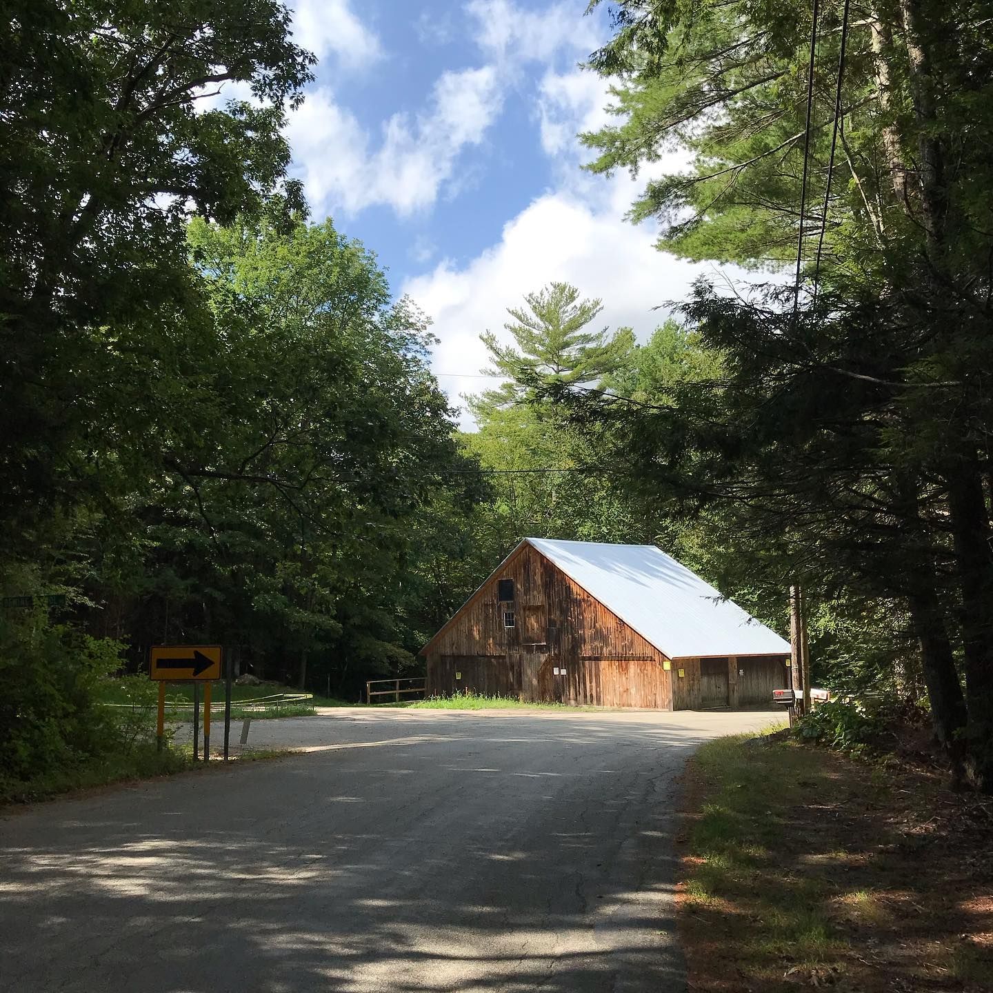 A rustic wooden barn sits at the end of a paved road, surrounded by trees under a blue sky.