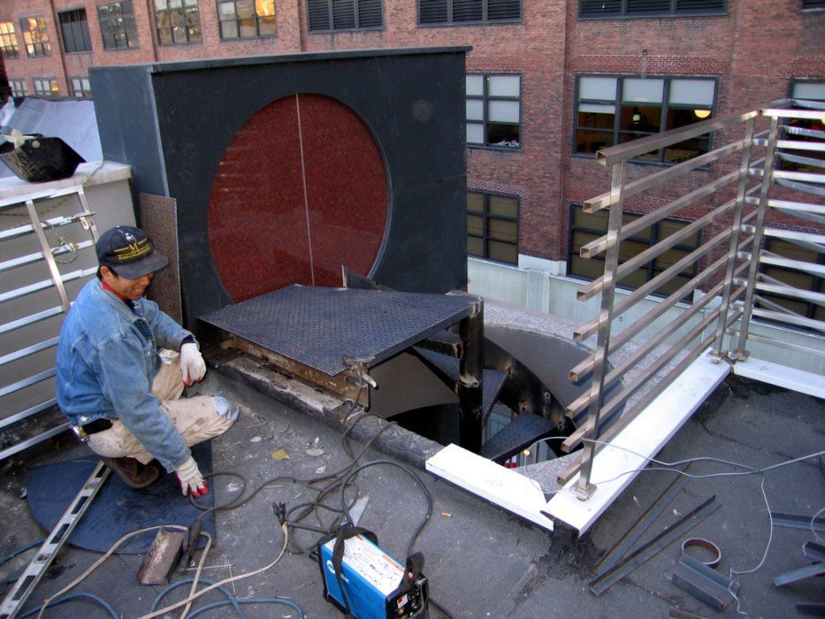 Construction worker on a rooftop, welding metal railings near a large vent.