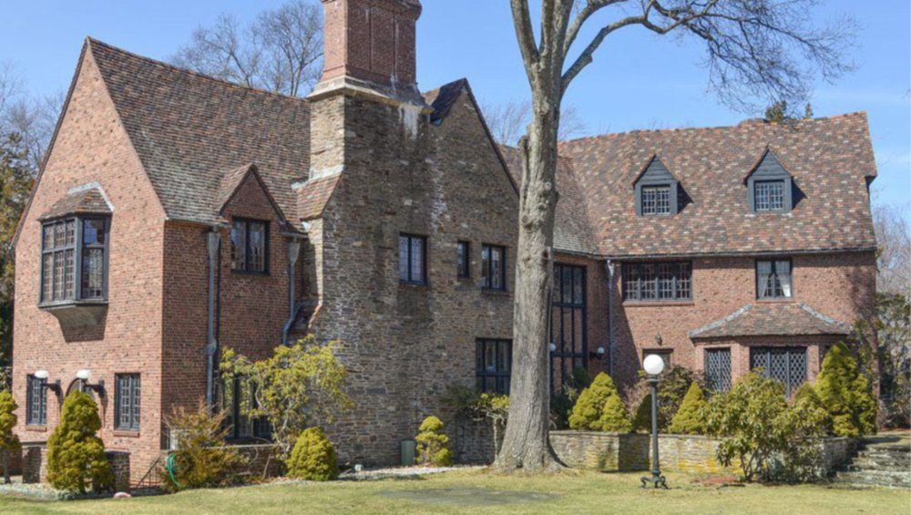 Brick house with multiple gables and a large chimney, set among trees on a grassy lawn.