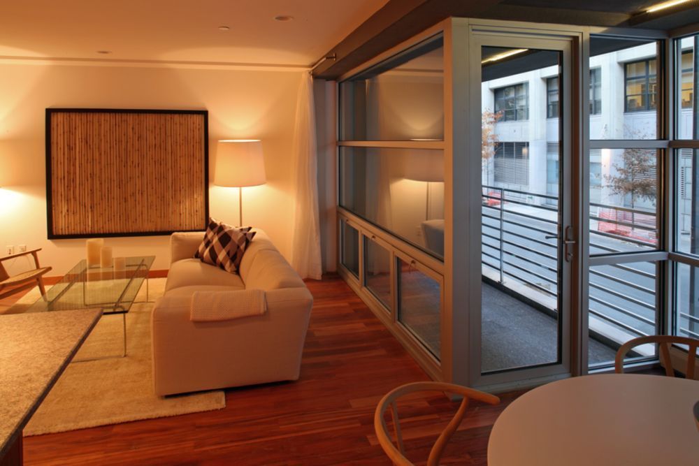 Living room with a glass-walled balcony. Beige sofa, artwork, and a glass coffee table. Wooden floor and natural light.