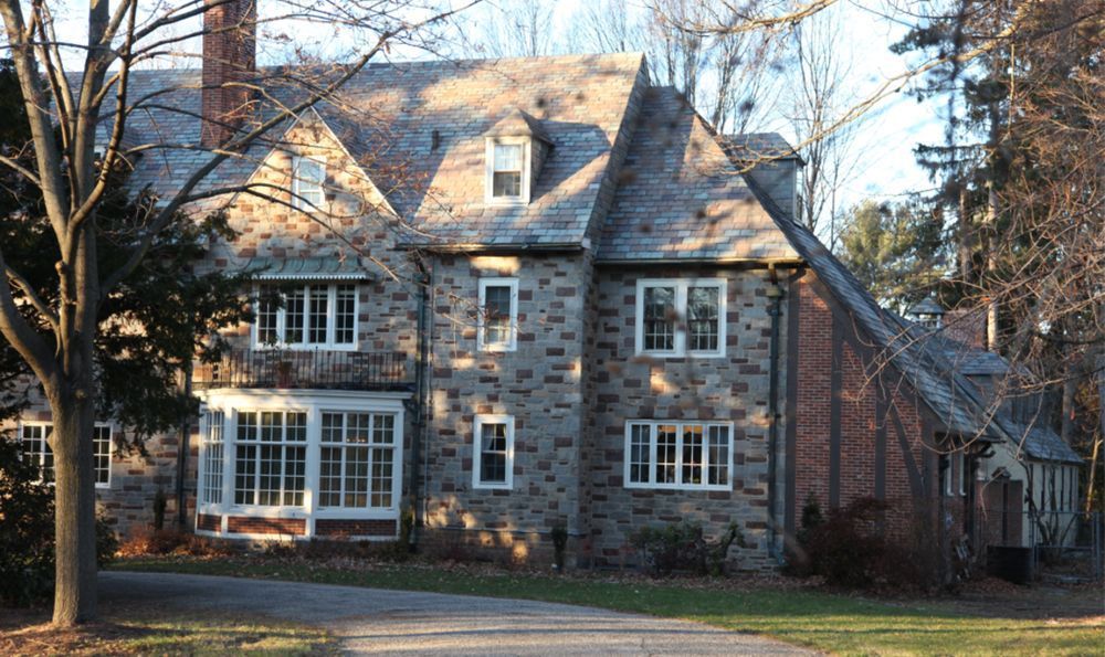 Stone-faced house with multiple gabled roofs, white-framed windows, and a driveway in a wooded setting.