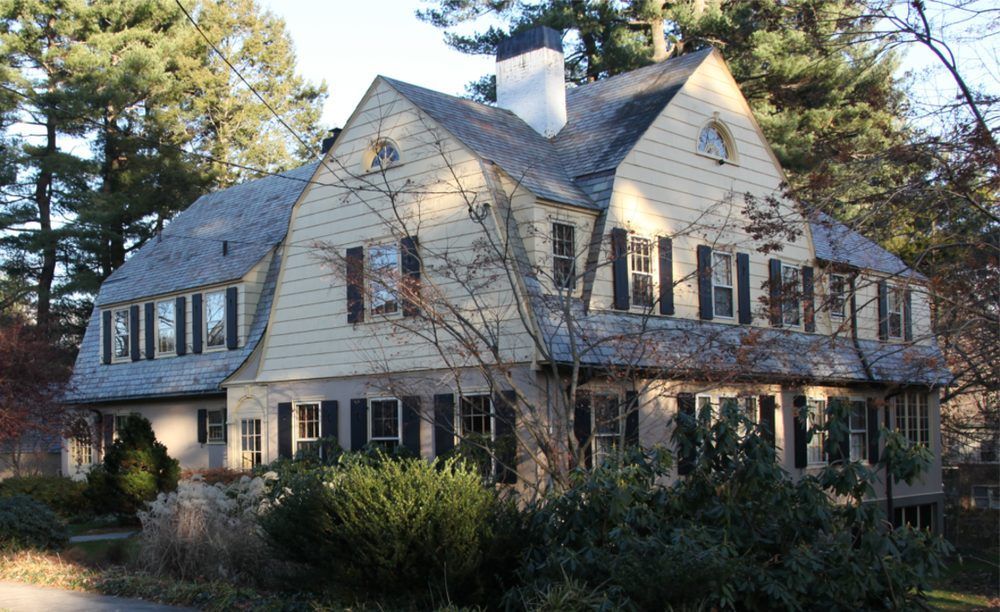 Two-story beige house with dark shutters and a slate roof, set among trees and shrubs.
