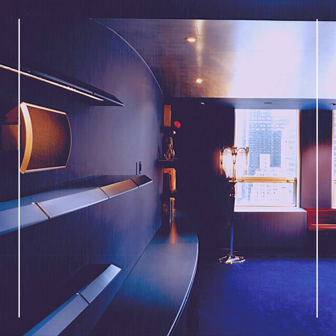 Dark blue, modern room with curved wall and city view through window. Shelves and gold accents.