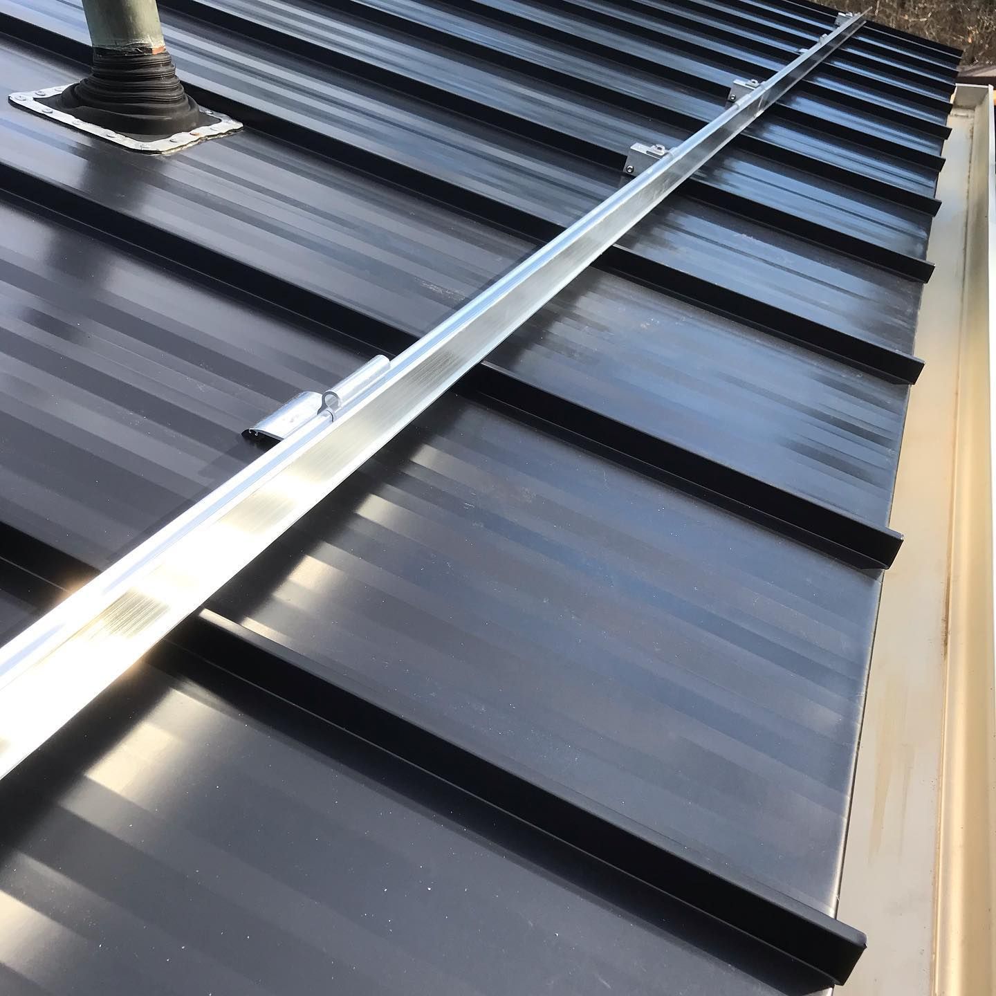 Black metal roof with a silver rail, near a beige gutter, and a vent pipe.