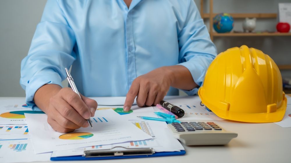 Man in blue shirt reviews charts, with yellow hard hat, calculator on desk.