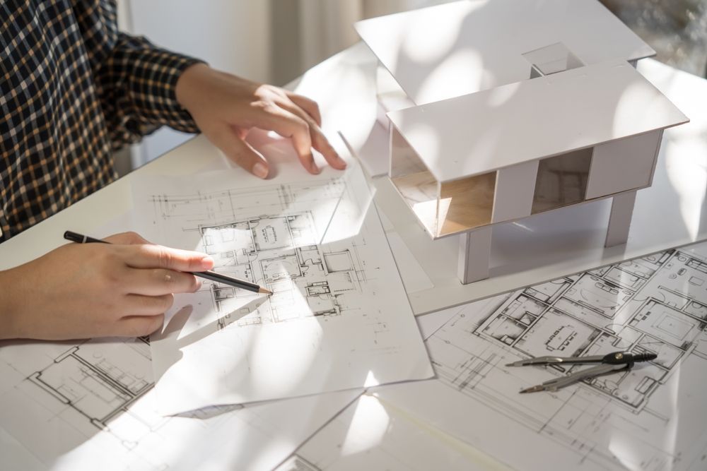 Person sketching architectural plans at a table with a model house and drafting tools.