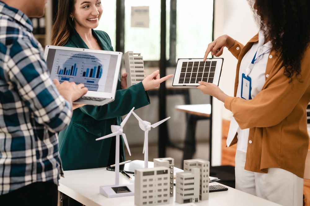 Three people discussing renewable energy models, including a solar panel, and a laptop with graphs.