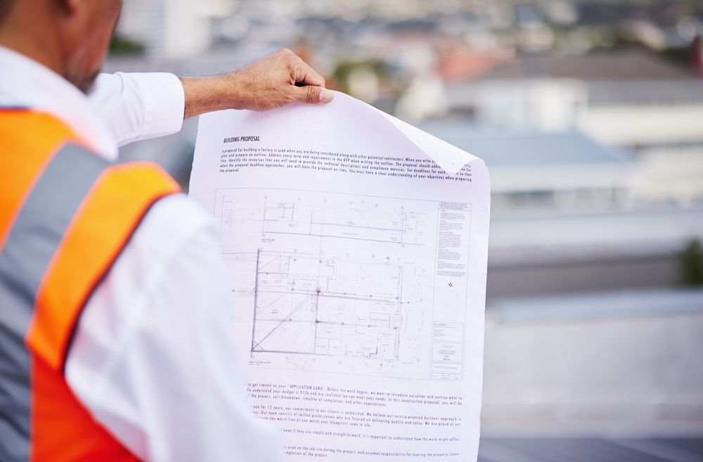 Construction worker wearing an orange vest, holding and reviewing blueprints on a rooftop.