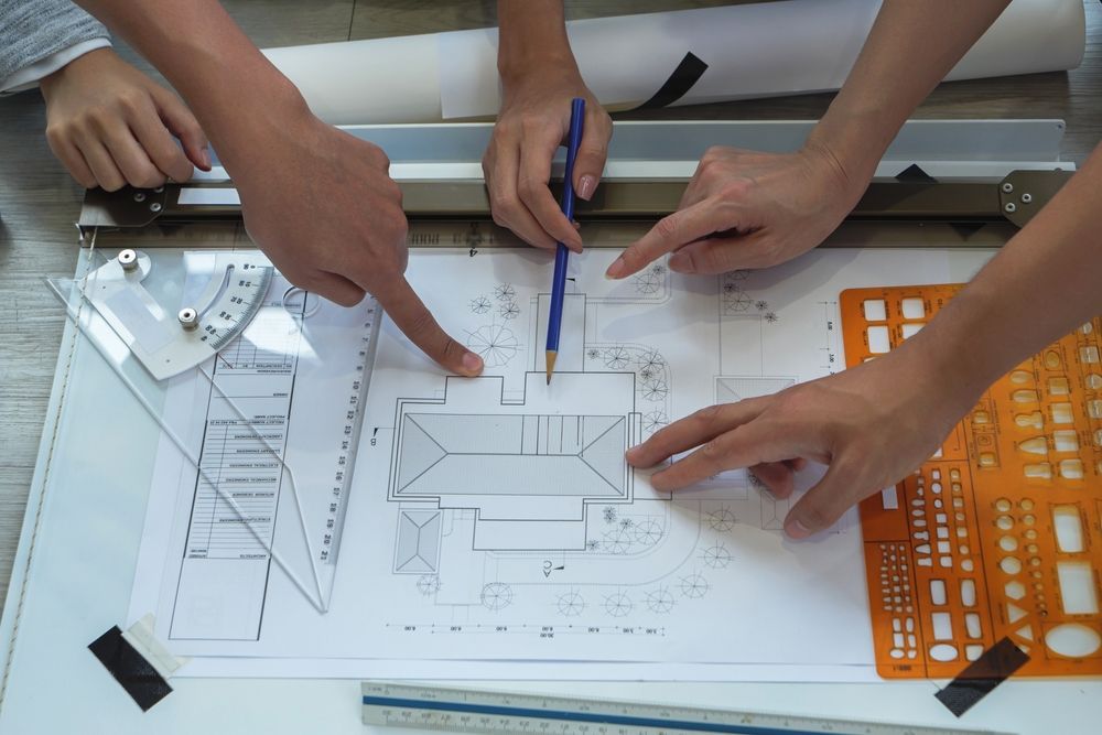 Hands pointing at architectural blueprints on a drafting table, with drawing tools.