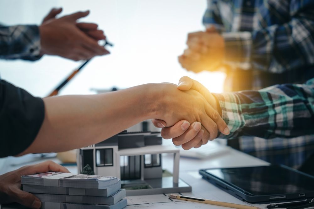 People shaking hands over a model house, money, and tablet on a table, with others in the background.