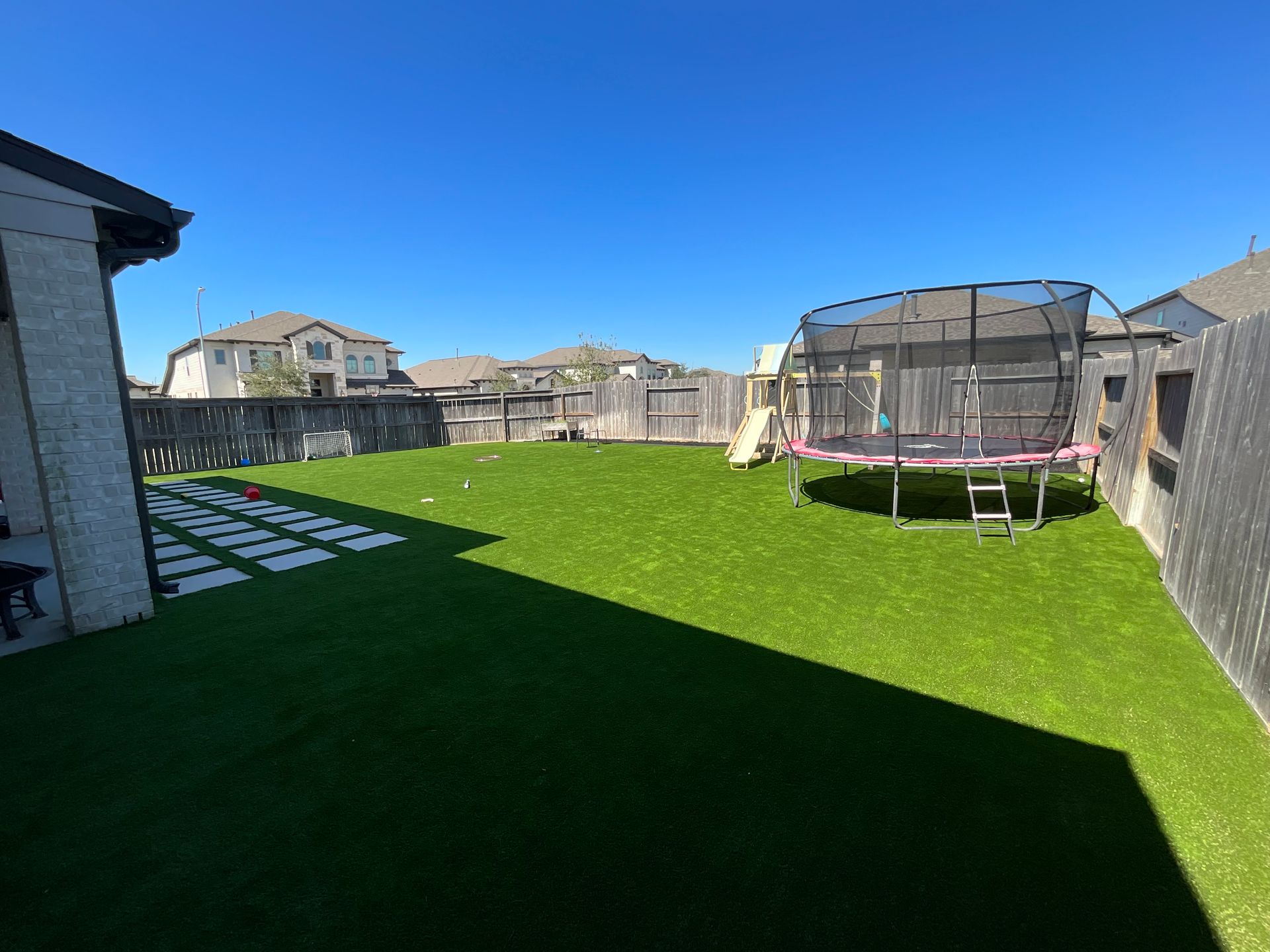 Sunny backyard with green artificial turf, trampolines, and houses under a clear blue sky