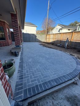 Brick-paved backyard patio with curved edge, potted plants, and fence under a clear blue sky