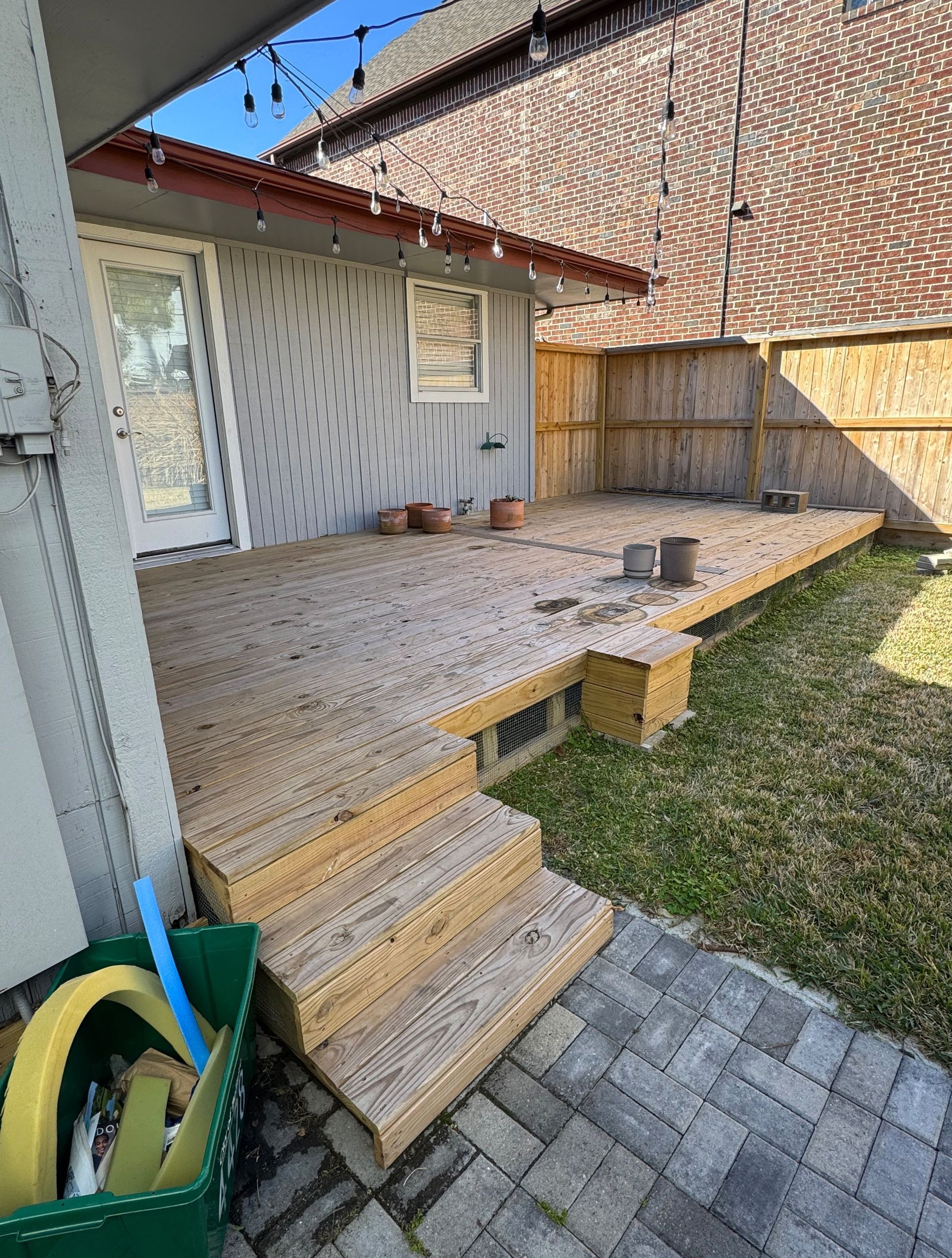 Wooden backyard deck with steps, railing, and potted plants beside a grassy yard and paved path