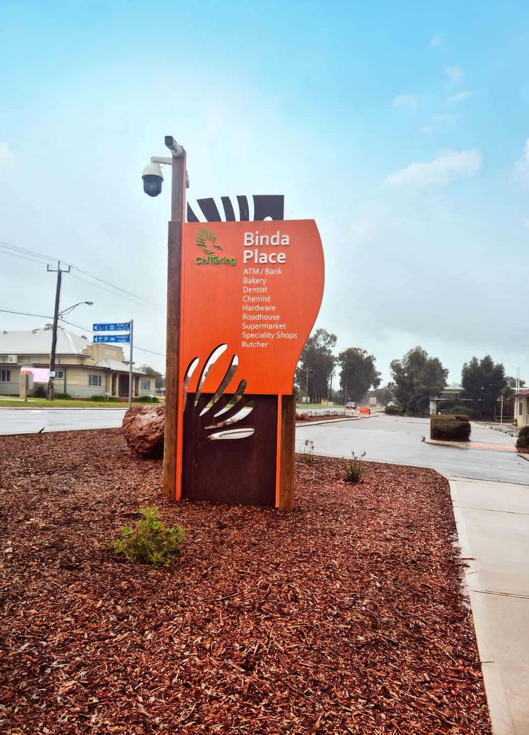 Sign for Birds Place community, orange and brown, with brown gravel bed, overcast sky.