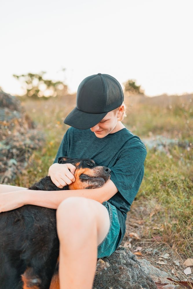 A person in a black cap cuddles a black and brown dog outdoors.