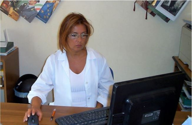 Donna in camice bianco alla scrivania, che guarda un computer. Ha un mouse in mano.