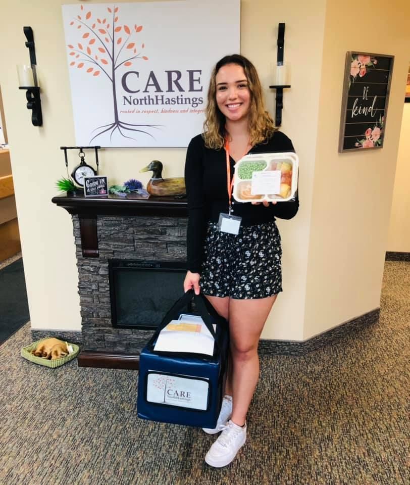 A woman is holding a box of food in front of a sign that says care north hastings.