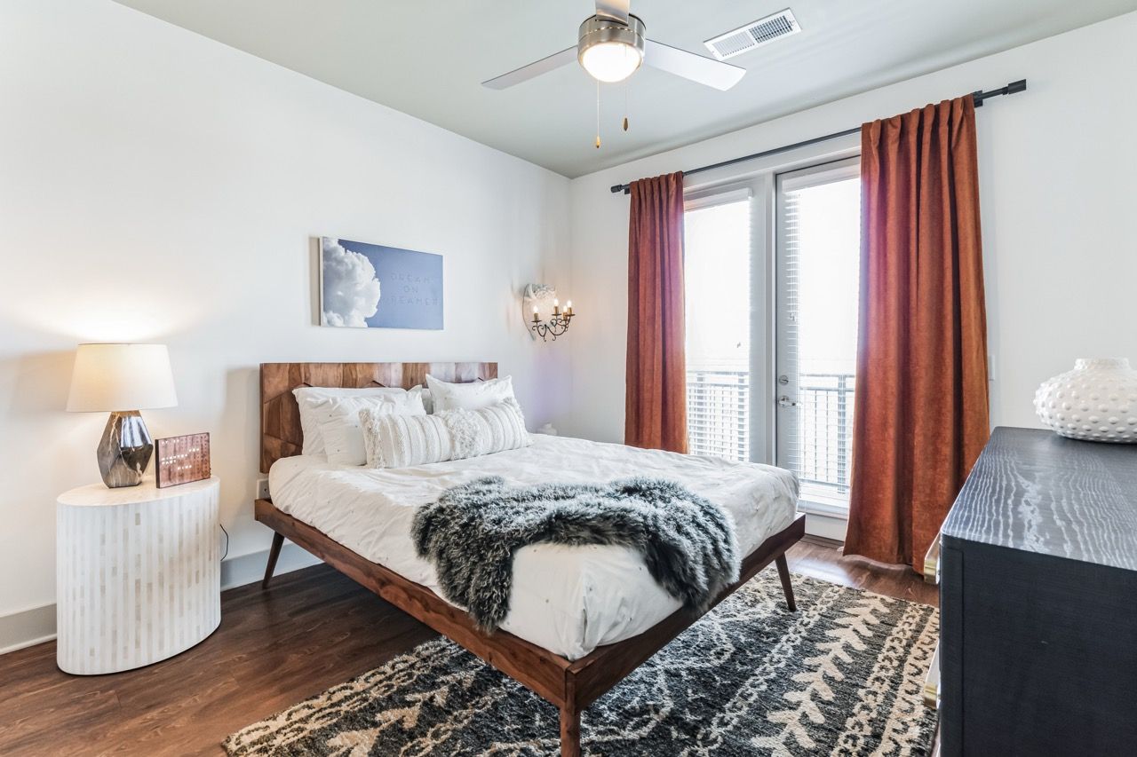 Bedroom in an apartment with a wooden platform bed, white bedding, and orange curtains beside a balcony door.