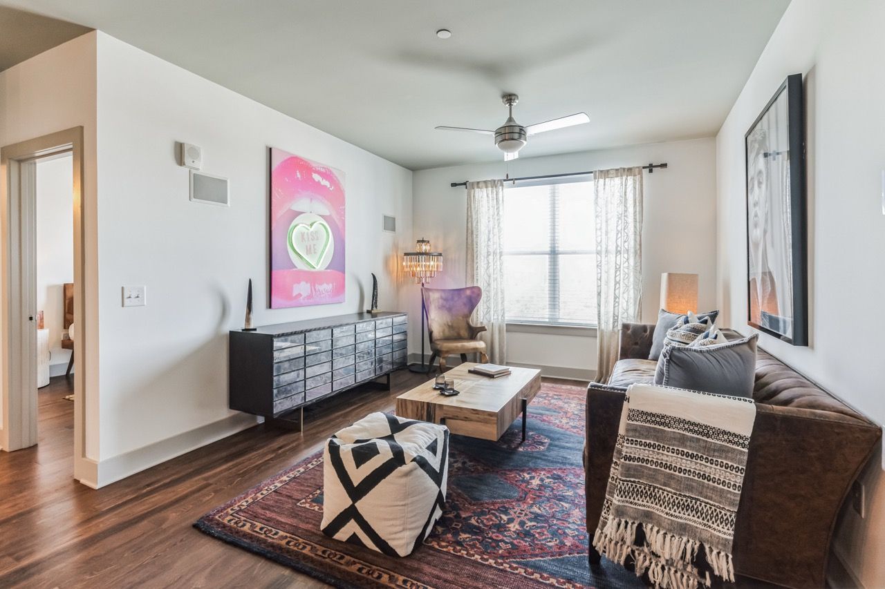 Living room in an apartment with a brown sofa, coffee table, colorful rug, and large window with curtains.