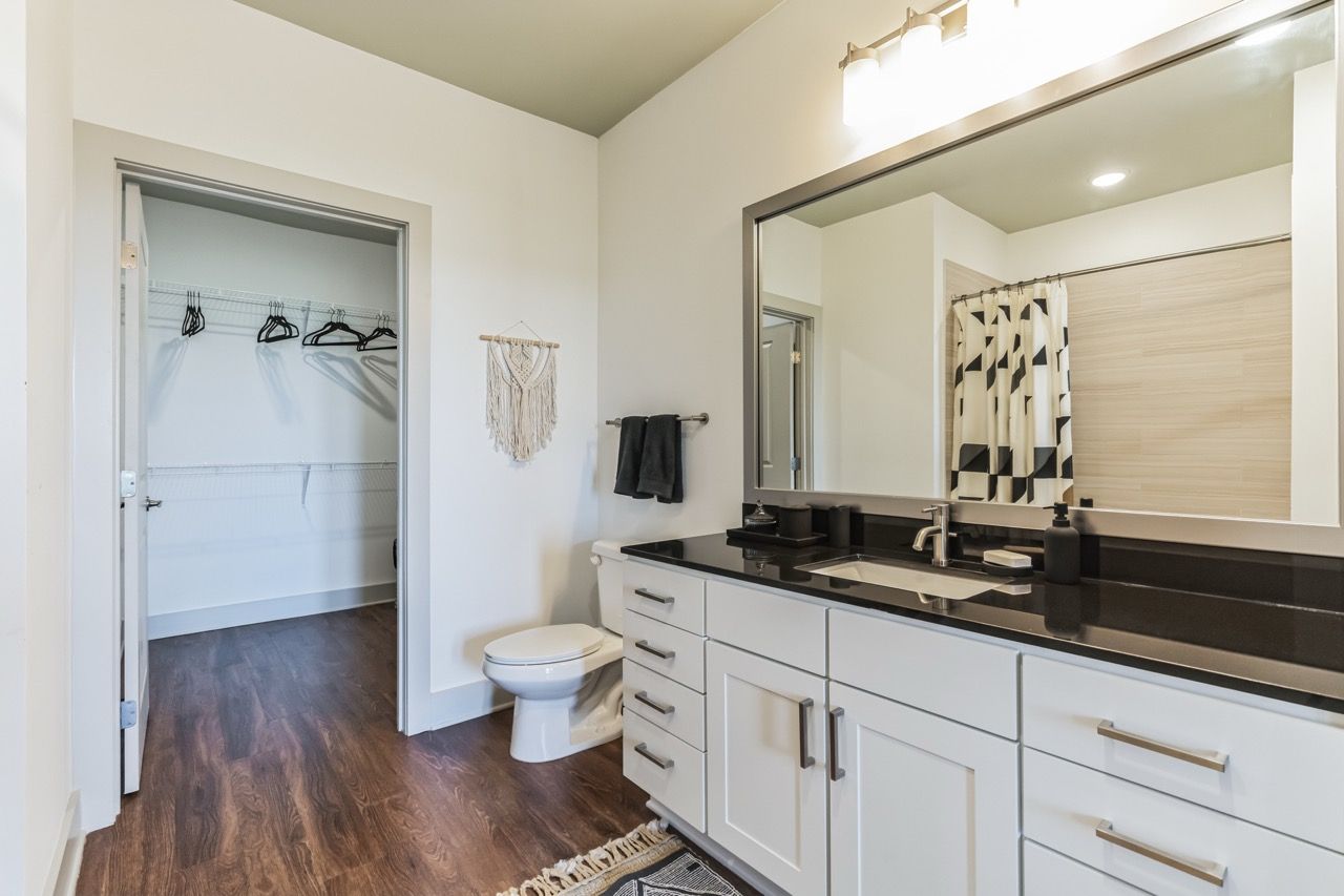 Bathroom with white cabinetry, black countertop, large mirror, toilet, and open walk-in closet.