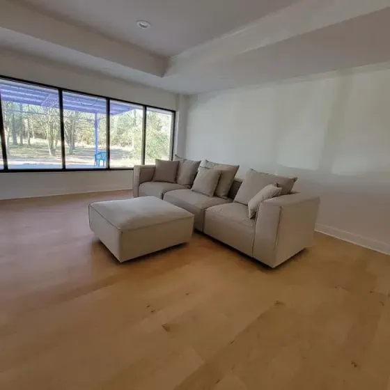 Modern living room with light-colored sofa and ottoman, large windows, and hardwood floors.
