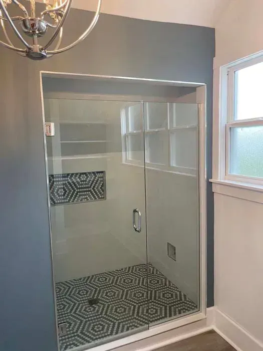 Glass shower with patterned tile floor and geometric tile accent on a white wall. Next to a frosted window.