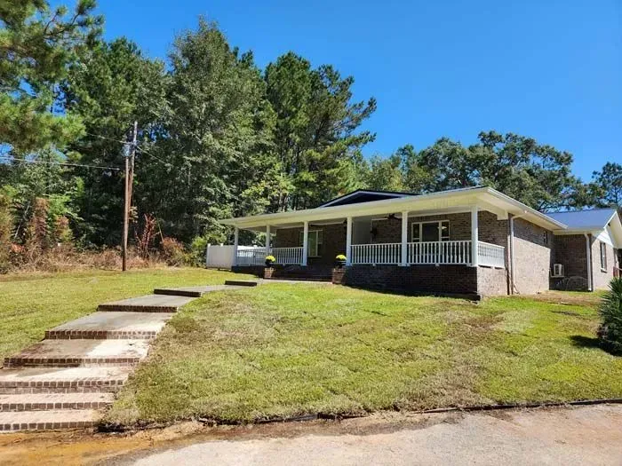 A single-story brick house with a porch and steps leading up from the yard, trees in the background.
