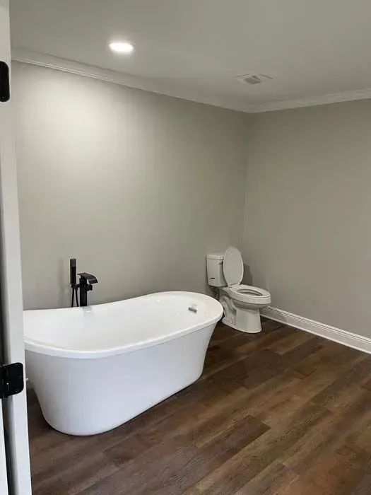 Freestanding white bathtub and toilet in a bathroom with wood flooring and light gray walls.