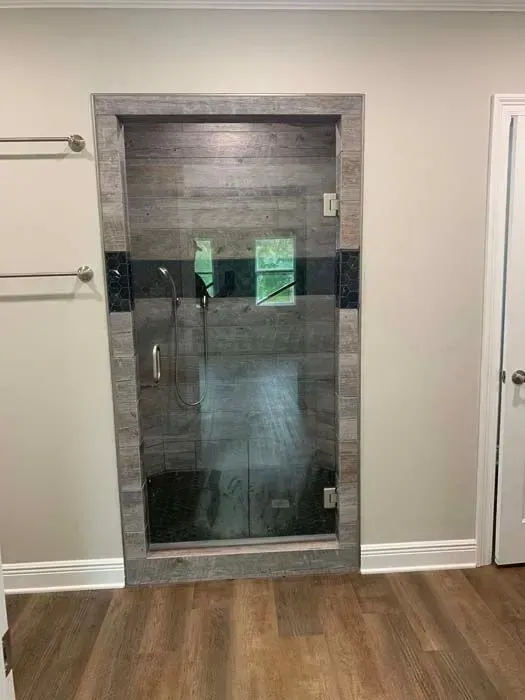 Shower with glass door in a bathroom, framed by wood-like gray tile. Adjacent to a door and towel rack.