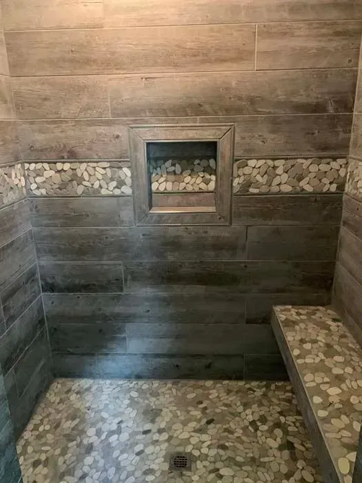 Shower with wood-look tile walls, stone-tiled floor, bench, and a recessed shelf.