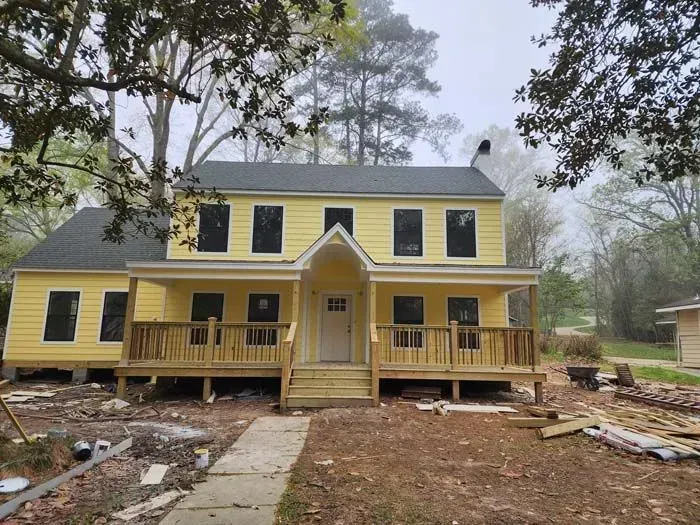 Yellow two-story house with dark windows, porch, and gray roof; cloudy day, surrounded by trees.