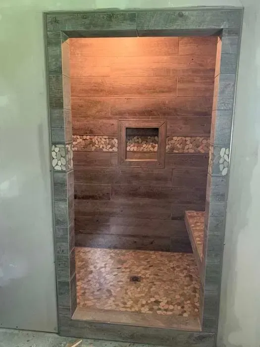 A walk-in shower with brown wood-look tiles, a pebble floor, and a built-in shelf.