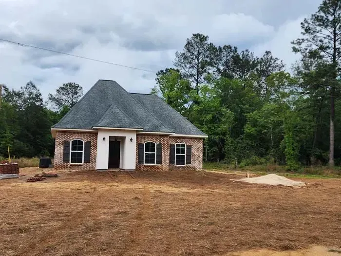 Newly constructed brick house with gray roof, white trim, and black shutters in a clearing, cloudy sky.