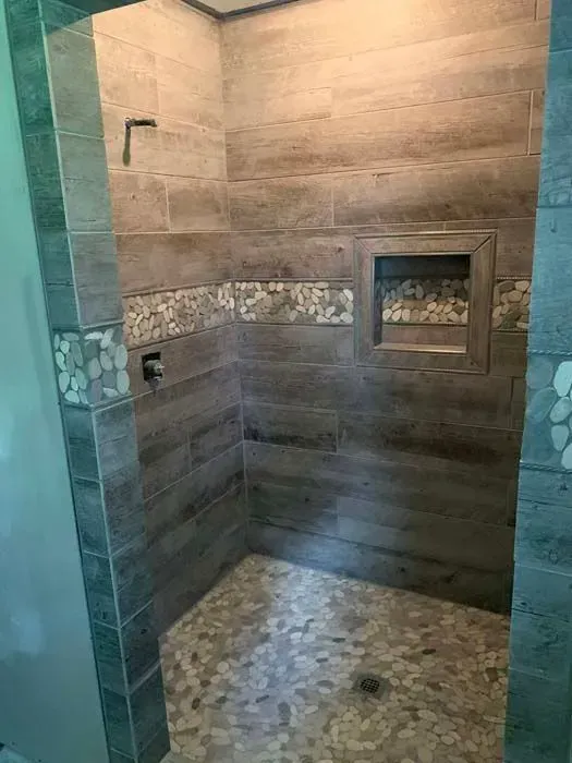 A newly renovated shower with wood-look tile walls, a pebble floor, and a decorative tile accent.