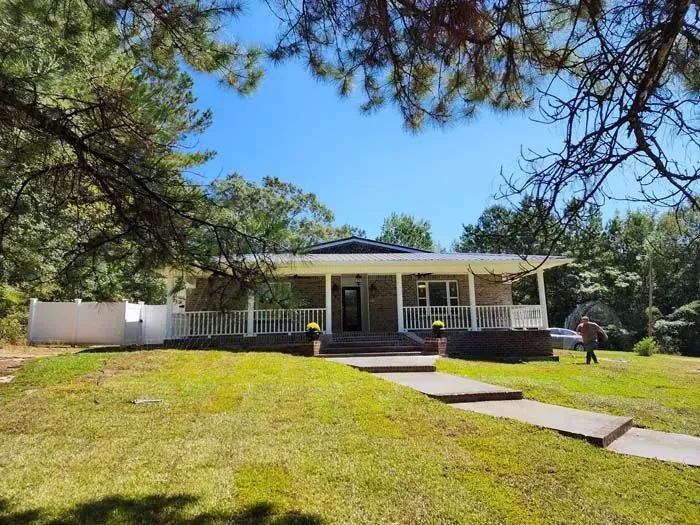 Ranch-style house with a covered porch, white railings, and brick facade, under a bright blue sky; person in the yard.